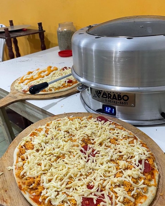 Duas pizzas montadas na bancada, ainda cruas, ao lado do forno marcando 121 graus